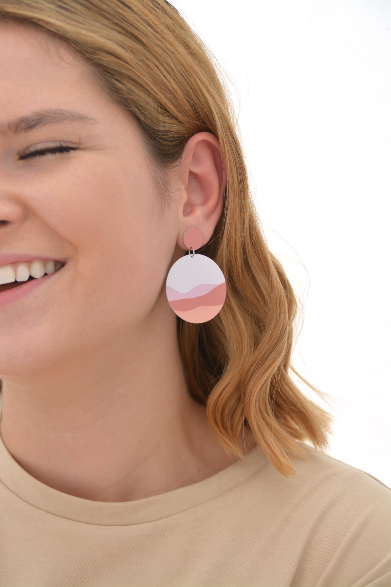 The image shows a woman wearing a Kitty Came Home large circle drop stud earring in her left ear. The top stud is 12 millimetres in diameter and the lower dangle circle is 36 millimetre in diameter. The design is waiting for the sun by Satin and Tat. Pale pink and orange forms create a landscape of rolling hills beneath a pale pink sky.