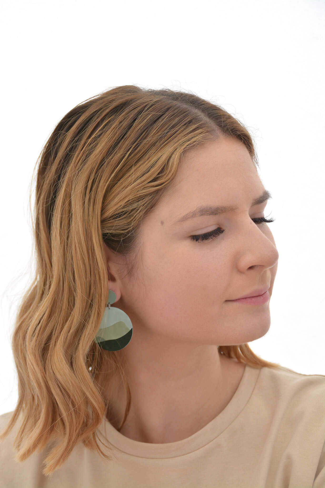 This is an image of a woman wearing a Kitty Came Home drop stud earring in her right ear. The top circle is 12 millimetres in diameter and the dangle circle is 36 millimetres in diameter. The design is called moondance by Satin and Tat. Darker green shapes form a landscape of rolling hills beneath of pale green night sky.