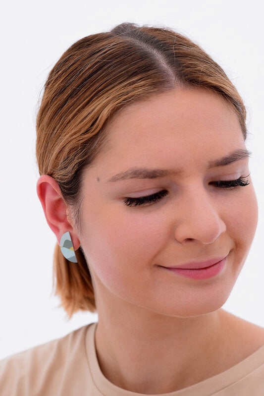 This image shows a woman wearing a Kitty Came Home stud earring in her right ear. The semi circle shape is 25 millimetres in diameter. The design is daylight drive by Satin and Tat. Green and yellow shapes form a landscape of gently rolling hills beneath a pale green sky.
