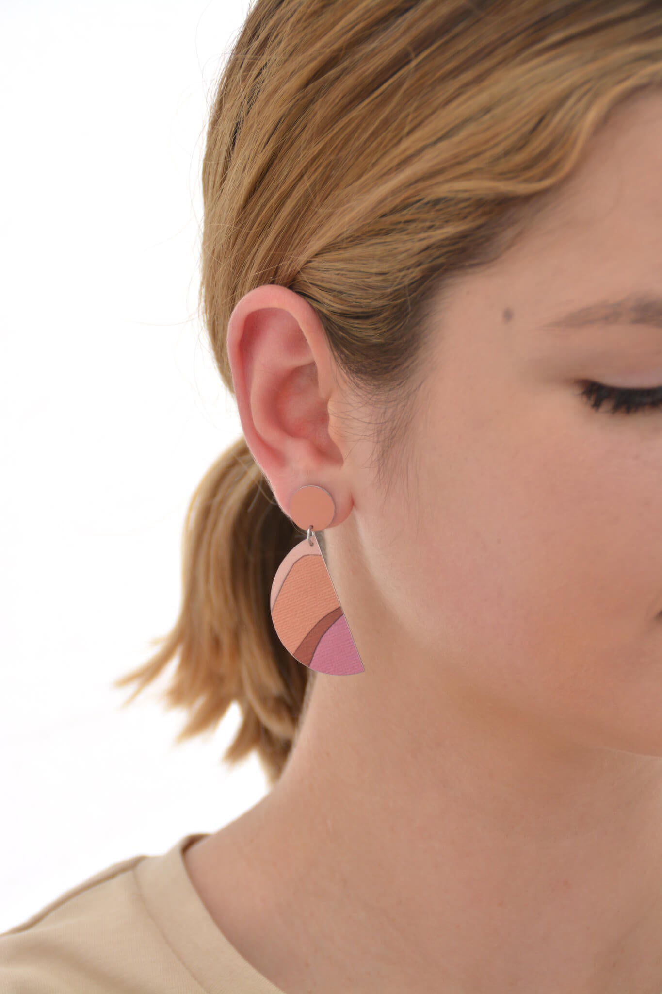 This is an image of a woman wearing a Kitty Came Home drop stud earring in her right ear. The top circle is 12 millimitres in diameter and the dangling semi circle beneath is 36 millimetres in diameter. The design is called the golden dawn by Satin and Tat. Burgundy, orange and terracotta shapes form a landscape of gently rolling hills beneath an orange-pink sky.