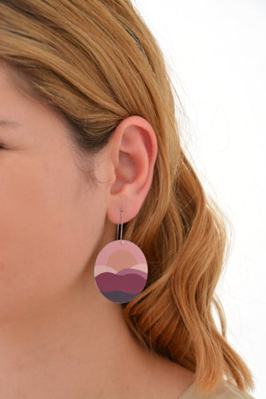 This is an image of a woman wearing a Kitty Came Home earring in her left ear. The flat circle is 36 millimetres in diameter and hands from a shepherds hook. The design is the night comes down by Satin and Tat. Magenta, burgundy and dusty pink shapes form an undulating landscape above which a golden sun sets in a terracotta sky.