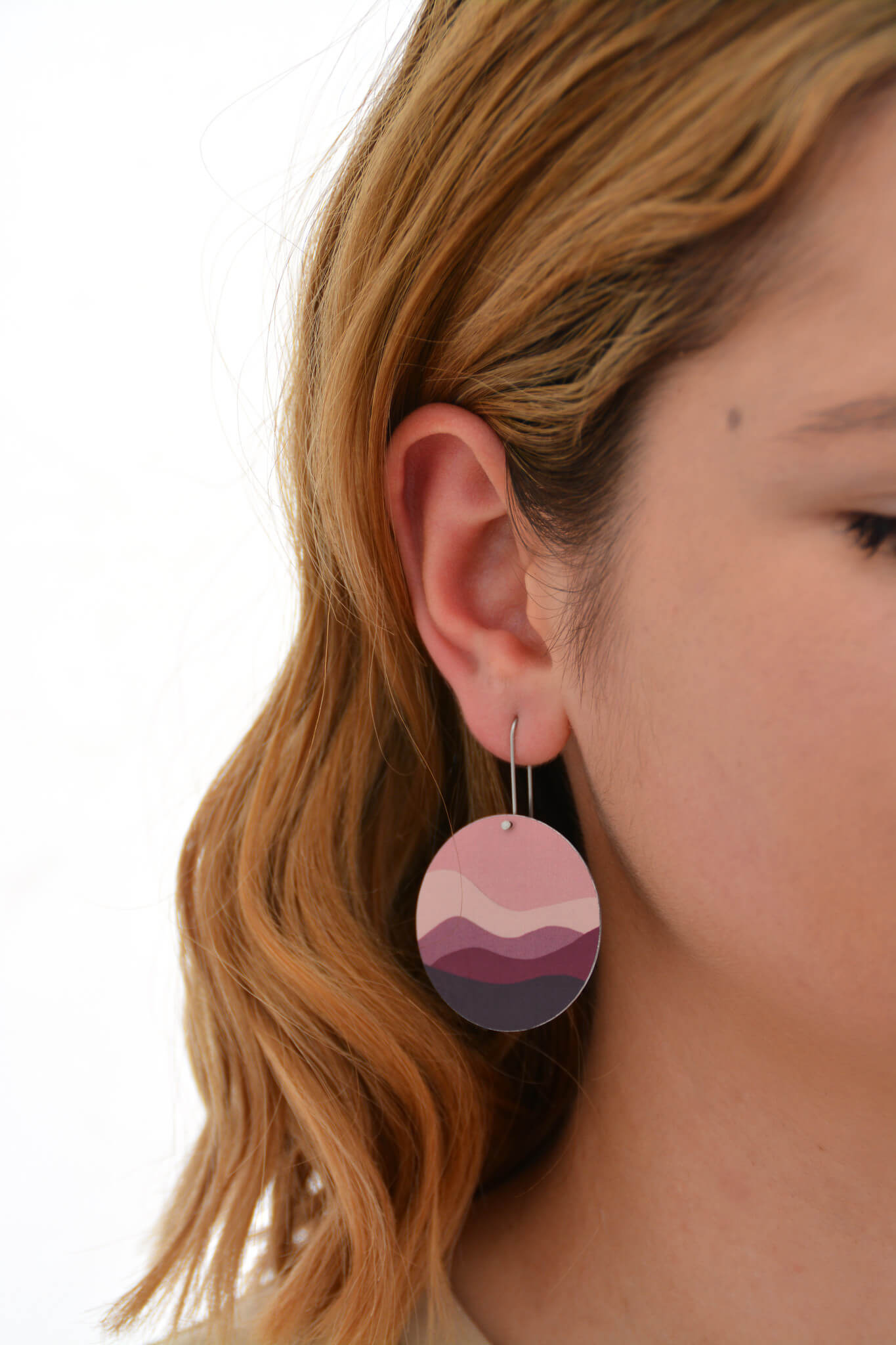 This is an image of a woman wearing a Kitty Came Home earring in her right ear. The flat circle is 36 millimetres in diameter and hands from a shepherds hook. The design is the night comes down by Satin and Tat. Magenta, burgundy and dusty pink shapes form an undulating landscape beneath a terracotta sky.