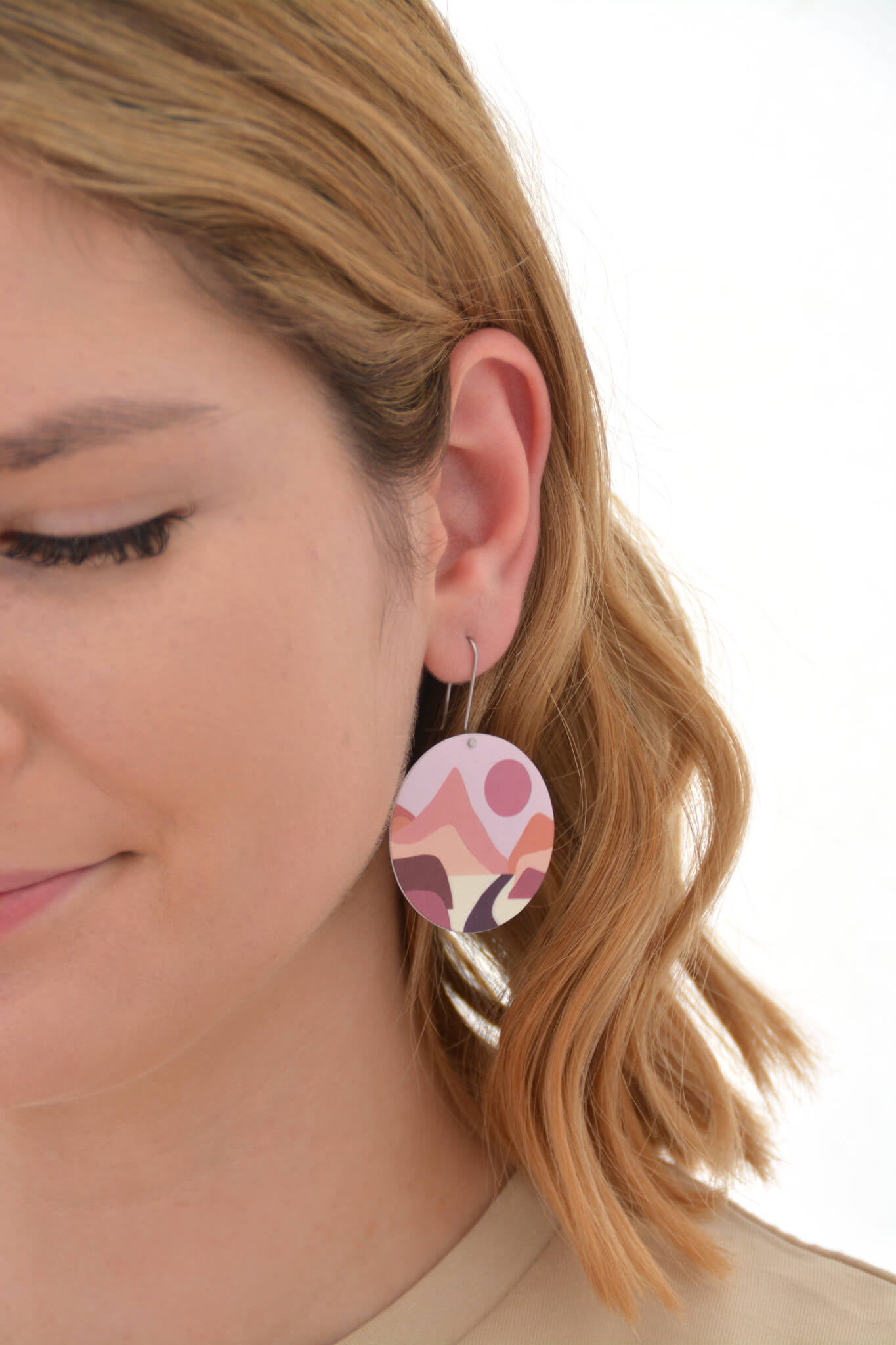 This is an image of a woman wearing a Kitty Came Home earring in her left ear. The flat circle is 36 millimetres in diameter and hangs from a shepherds hook. The design is lily of the valley by Satin and Tat. A terracotta coloured sinks in a pale pink sky towards a landscape of magenta, burgundy and pink hills. A path proceeds across the valley floor towards a cleft in the far mountain range.