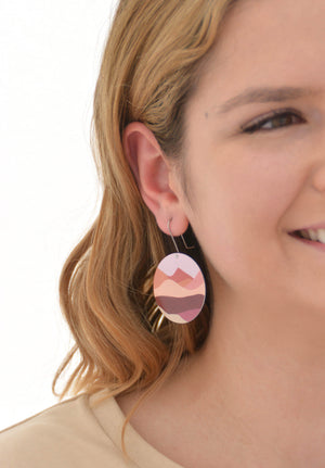 This is an image of a woman wearing a Kitty Came Home earring in her left ear. The flat circle is 36 millimetres in diameter and hangs from a shepherds hook. The design is lily of the valley by Satin and Tat. A landscape of magenta, burgundy and dusty pink hills lies beneath a pale pink sky. 