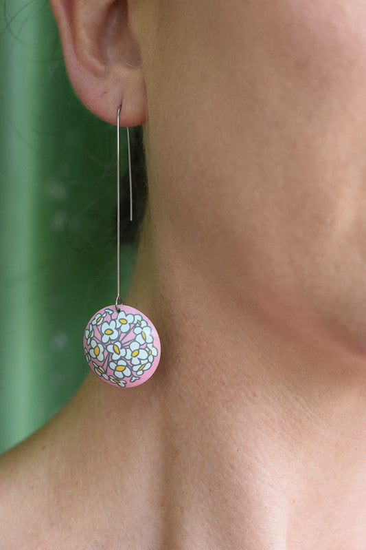 Photo of an earring on a model. The earring is a 26 millimetre in diameter domed circle on a 50 millimetre long stainless steel hand formed hook. The earrings are aluminium and the printed design is a Sweet Alyssum flower on a soft pink background. The design is printed on the convex side of the dome.