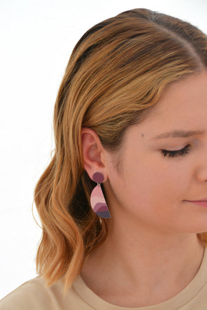 This is an image of a woman wearing a Kitty Came Home drop stud earring in her right ear. The top circle is 12 millimitres in diameter and the dangling semi circle beneath is 36 millimetres in diameter. The design is called the night comes down by Satin and Tat. Magenta and burgundy shapes form a landscape of gently rolling hills beneath a dusty pink sky.