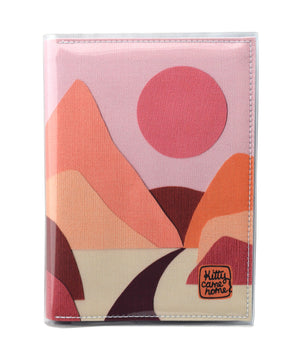 This is an image of the front of a Kitty Came Home A5 journal in the 'Lilly of the valley' design by Satin and Tat. A pink sun in a dusty pink sky above a landscape of orange, pink, burgundy and magenta hills. A path leads between the hills toward the distant mountains.