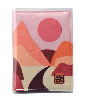This is an image of the front of a Kitty Came Home A6 journal in the 'Lilly of the valley' design by Satin and Tat. A pink sun in a dusty pink sky above a landscape of orange, pink, burgundy and magenta hills. A path leads between the hills toward the distant mountains.