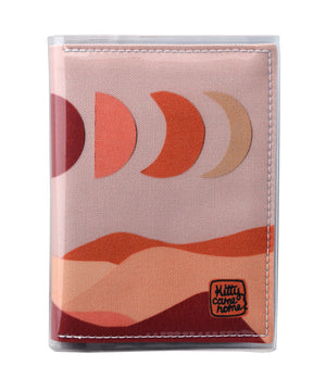 This is an image of the front of a Kitty Came Home A6 journal in the 'Moonshadow drive’ design by Satin and Tat. The moon appears in all its phases in a dusty pink sky above a landscape lit by the moon's colours: burgundy, oranges, and apricot.