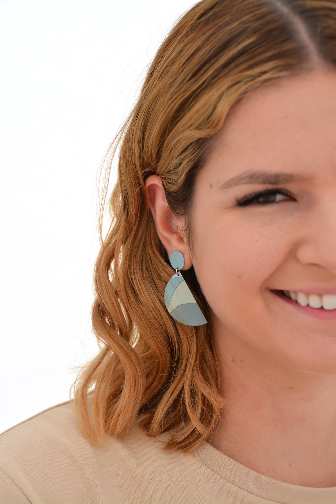 The image shows a woman wearing a Kitty Came Home double drop stud semi-circle earring in her right ear. The top stud is a 12 millimetre diameter circle, and the lower dangle is a 36 millimetre diameter semi-circle. The design is the night grows pale by Satin and Tat. Pale blue and green forms create a landscape of gently rolling hills.