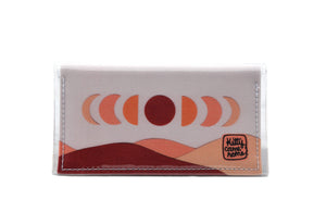 This is an image of the front of a Kitty Came Home bifold mini purse clutch in 'The same moon shines' design by Satin and Tat. The moon appears in all its phases in a white sky above a landscape lit by the moon's colours: burgundy, oranges and apricot. This is the small size.